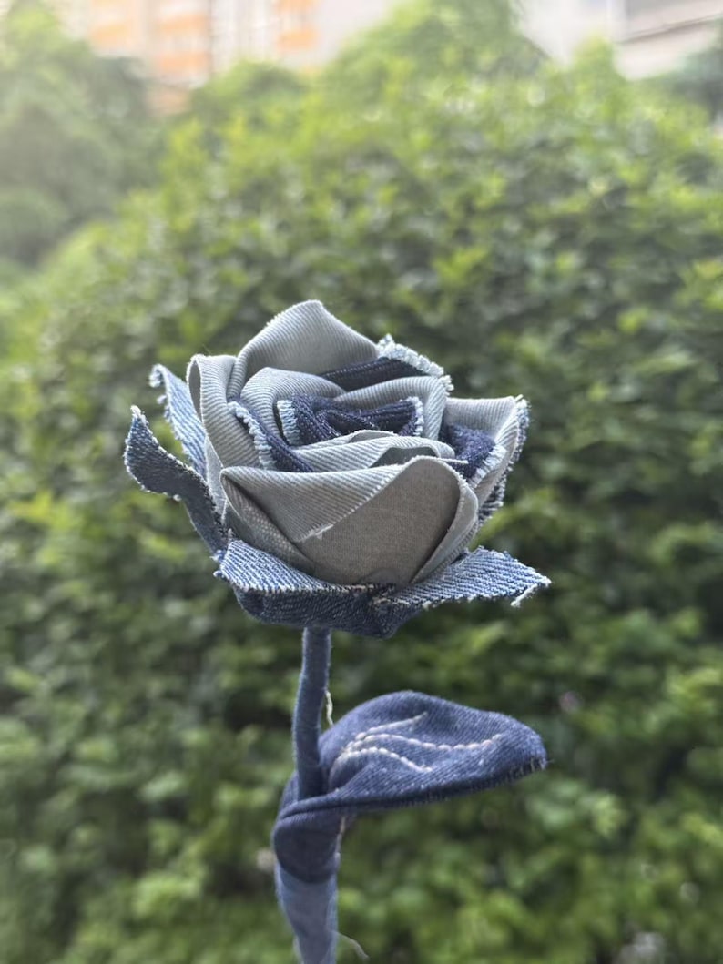 Pode incluir: Uma rosa de ganga artesanal com caule e folha. As p&eacute;talas da flor s&atilde;o feitas de v&aacute;rias tonalidades de ganga azul, criando uma apar&ecirc;ncia texturizada e &uacute;nica. O caule e a folha tamb&eacute;m s&atilde;o feitos de ganga.
