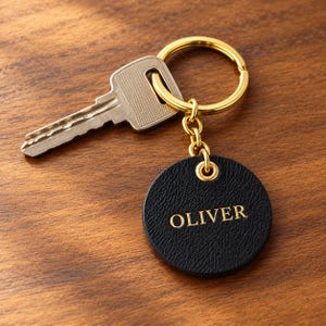 May include: A gold-toned metal keyring attached to a silver-toned house key and a circular black textured tag. The tag features the name OLIVER embossed in gold lettering. The keyring includes a small gold chain link connecting the tag to the ring.