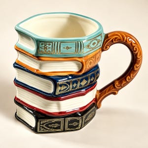 Può includere: Una tazza in ceramica a forma di pila di cinque libri con un manico marrone curvo e decorato. I libri presentano dorsi color ottanio, arancione, blu scuro, rosso e nero, ciascuno decorato con motivi dorati e bordi delle pagine bianchi.