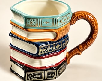 Op de afbeelding: Een keramische mok in de vorm van een stapel van vijf boeken met een gebogen, sierlijk bruin handvat. De boeken hebben ruggen in de kleuren groenblauw, oranje, donkerblauw, rood en zwart, elk versierd met gouden patronen en witte paginaranden.