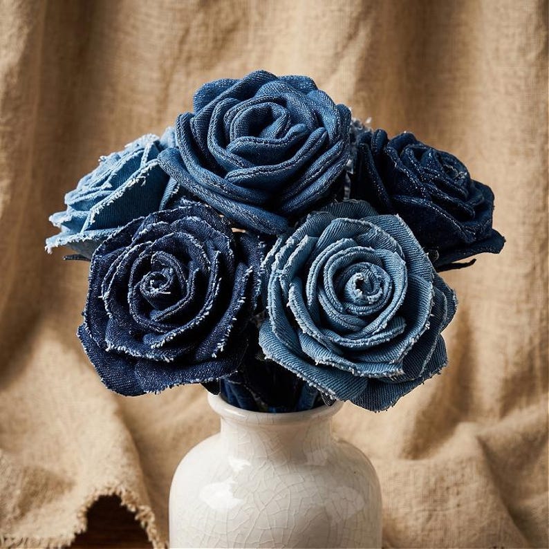 Pode incluir: Um buqu&ecirc; de rosas de ganga azul, dispostas em um vaso de cer&acirc;mica branca. As rosas t&ecirc;m um detalhe de borda desfiada, e o vaso tem um acabamento craquelado. O fundo &eacute; um tecido bege neutro.