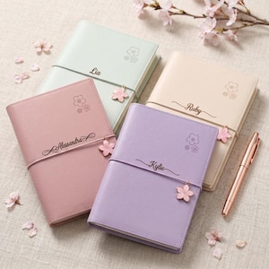 Op de afbeelding: Vier gepersonaliseerde agenda's in pastelkleuren: lichtgroen, roze, crème en lavendel. Elke agenda heeft een naam, een bloemmotief en een roze bloemcharm. Een roségouden pen ligt naast de agenda's.