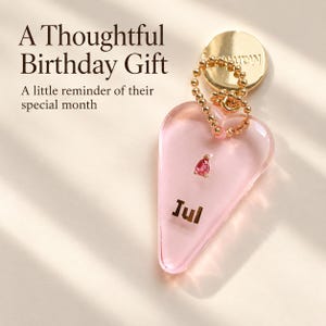 May include: A heart-shaped pink acrylic charm featuring the text "Jul" and a small pink teardrop-shaped gemstone. The charm is attached to a gold-coloured ball chain and a circular gold-coloured metal tag. Text on the image reads "A Thoughtful Birthday Gift" and "A little reminder of their special month."