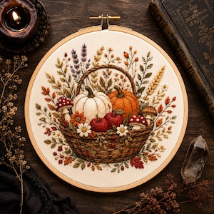 May include: An embroidery hoop containing a fabric panel with a detailed needlework design of a woven basket filled with two pumpkins, two red apples, and two red-and-white spotted mushrooms, surrounded by various autumn flowers and wheat stalks.