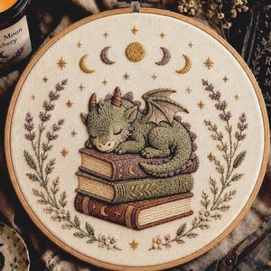以下が含まれることがあります： 3冊の本の山の上で眠る緑色のドラゴンの刺繍が施された刺繍枠。デザインには、両サイドに刺繍されたラベンダーの枝、ドラゴンの上の月相サイクル、クリーム色の布地に散りばめられた小さな星が含まれています。