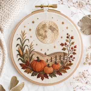 May include: A circular wooden embroidery hoop containing a fabric panel with a hand-stitched autumn scene. The design features a large textured moon, three orange pumpkins, stalks of corn, a wooden fence, and fall foliage in shades of brown.