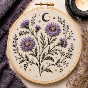 Puede incluir: Un bastidor de bordado circular de madera que contiene un panel de tela con flores moradas similares a margaritas, tallos y hojas verdes bordadas a mano. El diseño incluye una luna creciente negra y pequeños detalles de estrellas doradas.