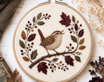 May include: A circular wooden embroidery hoop containing a fabric panel with a hand-stitched design of a small brown wren perched on a branch. The bird is surrounded by an intricate wreath of embroidered autumn leaves in shades of burgundy, olive green, and tan, accented with small dark brown berries.