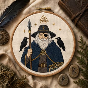 May include: An embroidery hoop featuring a stitched portrait of a bearded figure wearing a wide-brimmed hat with a rune, an eye patch, and a dark blue cloak. The figure holds a spear, flanked by two black ravens, with a Valknut symbol above.