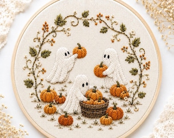 May include: A circular wooden embroidery hoop containing a fabric panel with a hand-stitched design of three white ghosts interacting with orange pumpkins. The scene is framed by a delicate embroidered vine with green leaves and small berries.