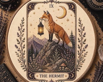 May include: A circular embroidery hoop featuring a fabric panel with a detailed illustration of a fox standing on a rocky mountain peak holding a lantern. The design includes a crescent moon, stars, pine trees, and the text THE HERMIT with Roman numeral IX.