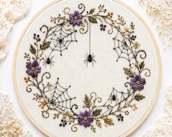 May include: A circular wooden embroidery hoop containing a fabric panel with a hand-stitched wreath design. The wreath features purple flowers, green leaves, small orange berries, and black spiderwebs with three small black spiders.
