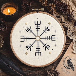 May include: A circular wooden embroidery hoop containing off-white fabric with a dark blue Vegvisir symbol stitched in the centre. The design features small gold and silver star accents and a gold-stitched ring at the very centre of the compass.