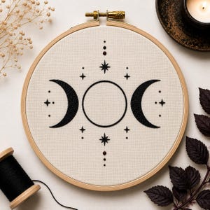 May include: A circular wooden embroidery hoop containing off-white fabric with a black stitched design of a central full moon flanked by two crescent moons, surrounded by small star and dot motifs arranged in a symmetrical vertical pattern.