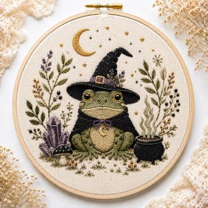 May include: A circular embroidery hoop containing a fabric panel with a stitched green frog wearing a black witch hat and cape. The design includes a crescent moon, stars, purple crystals, a small mushroom, and a black cauldron with smoke.