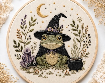 May include: A circular embroidery hoop containing a fabric panel with a stitched green frog wearing a black witch hat and cape. The design includes a crescent moon, stars, purple crystals, a small mushroom, and a black cauldron with smoke.