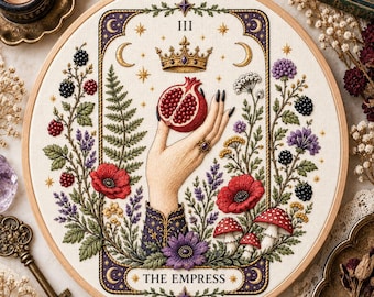 May include: A circular embroidery hoop featuring a tarot card design titled The Empress. The artwork depicts a hand holding a halved pomegranate beneath a crown, surrounded by detailed botanical elements including red poppies, blackberries, and mushrooms.