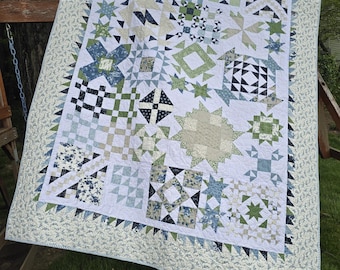 May include: A handmade quilt with a white background and various geometric patterns in shades of blue, green, and cream. The quilt features star, square, and floral designs, with a patterned border. The quilt is displayed outdoors.