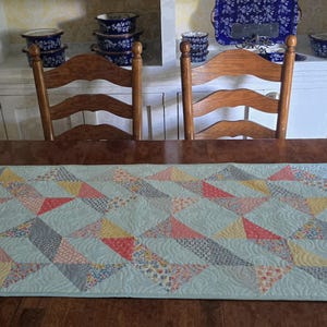May include: A light blue quilted table runner with a geometric pattern of triangles in shades of red, yellow, blue, and gray. The runner is placed on a dark brown wooden table, with two wooden chairs and a white cabinet in the background.