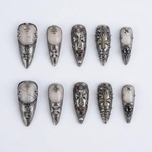 Può includere: Una collezione di dieci protezioni per unghie argentate. Ogni protezione presenta disegni intricati, tra cui croci, teschi e abbellimenti decorativi. Alcune hanno accenti di pietra colorata. Le protezioni sono disposte su una superficie bianca.