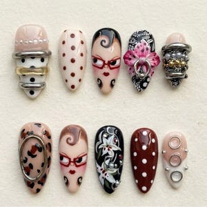 Op de afbeelding: Een verzameling van tien amandelvormige, versierde kunstnagels. Ontwerpen omvatten stippen, gezichten met brillen, bloempatronen en metalen accenten. Kleuren vari&euml;ren van nude en roze tot zwart en bruin.