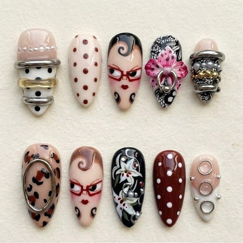 Op de afbeelding: Een verzameling van tien amandelvormige, versierde kunstnagels. Ontwerpen omvatten stippen, gezichten met brillen, bloempatronen en metalen accenten. Kleuren vari&euml;ren van nude en roze tot zwart en bruin.