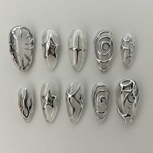 May include: A set of ten silver-toned, sculpted nail art designs. Each nail features a clear base with a unique metallic embellishment, including swirls, stars, and abstract shapes. The nails are oval-shaped and arranged in two rows.