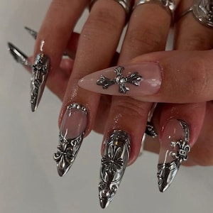 May include: Silver nail art featuring ornate metal designs. The nails are long and pointed, with a mix of cross and fleur-de-lis embellishments. Some nails have a clear base with silver accents, while others are fully silver.
