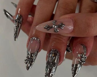 Puede incluir: Uñas plateadas con diseños metálicos ornamentados. Las uñas son largas y puntiagudas, con una mezcla de cruces y adornos de flor de lis. Algunas uñas tienen una base transparente con detalles plateados, otras son totalmente plateadas.