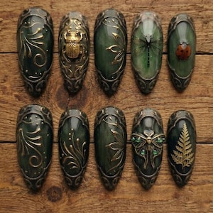 May include: A set of ten almond-shaped press-on nails featuring a dark green base with metallic gold accents. Designs include gold filigree swirls, a raised gold scarab beetle, a dragonfly, a ladybug, a moth with green gems, and a fern leaf.