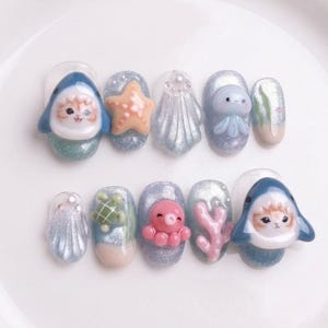 May include: A set of ten artificial nails with a marine theme. Each nail features a different 3D design, including a shark, starfish, seashell, jellyfish, and octopus. The nails are painted in shades of blue, pink, and white, with glitter accents.