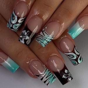 Może przedstawiać: Długie, kwadratowe sztuczne paznokcie z bazą typu french manicure i różnorodnymi wzorami. Niektóre paznokcie mają ciemnobrązowo-turkusowy wzór wirów, inne biały motyw kwiatowy, a jeszcze inne turkusowe paski zebry z małymi srebrnymi cyrkoniami.