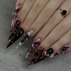 May include: Long stiletto-shaped artificial nails featuring a mix of glossy black, white, and deep red marbled polish. The nails are decorated with silver-toned metal charms including crosses, handcuffs, and small spikes, plus a heart-shaped ring.