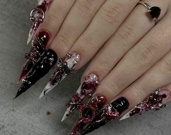 May include: Long stiletto-shaped artificial nails featuring a mix of glossy black, white, and deep red marbled polish. The nails are decorated with silver-toned metal charms including crosses, handcuffs, and small spikes, plus a heart-shaped ring.