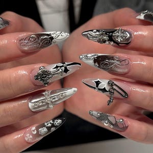 May include: Long almond-shaped artificial nails featuring a black and white colour palette. Designs include gothic calligraphy text, silver metallic cross charms, black star accents, a silver safety pin charm, and small clear rhinestones on a glossy finish.
