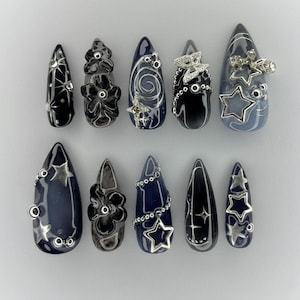 May include: A set of ten long, pointed artificial nails. The nails are painted in shades of black and navy blue, with silver embellishments including stars, flowers, and swirls. Some nails have a matte finish, while others are glossy.