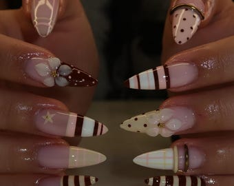 May include: Stiletto-shaped nails featuring a variety of designs including brown and white stripes, pink and white plaid patterns, brown polka dots, and three-dimensional white floral accents with gold centres, accented by small gold rings on two nails.
