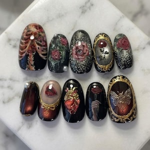 May include: A set of ten artificial nails with gothic-inspired art. Designs include a human rib cage, roses, an eye, a heart, and a moth. The nails are painted in black, red, gold, and silver, with a glossy finish.