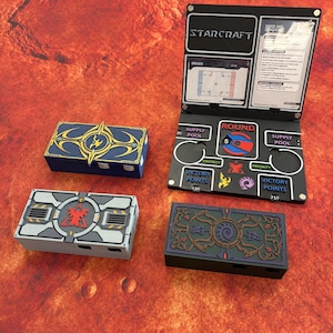 May include: A Starcraft board game accessory set featuring a black folding dashboard with game text, round and victory point trackers, and three rectangular storage boxes in blue, grey, and black with distinct faction-themed graphic designs.