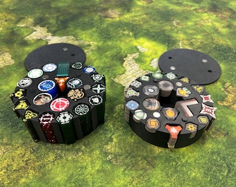 May include: Two black, circular game token holders with various colorful tokens. Each holder has a removable lid. The tokens feature different designs and colors, including red, green, blue, and yellow. The holders are on a green surface.