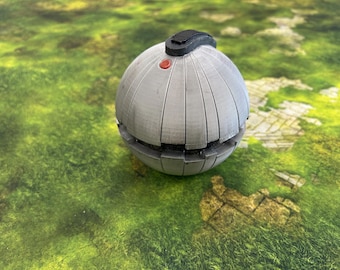 May include: A gray, spherical object with vertical lines and a black top. A small red button is visible on the top. The object is resting on a green and brown surface.