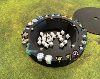 May include: A black, circular dice tray with a central well containing white dice with red markings. The tray has a series of decorative tokens around the edge. A black lid is on the right side. The tray sits on a green surface.