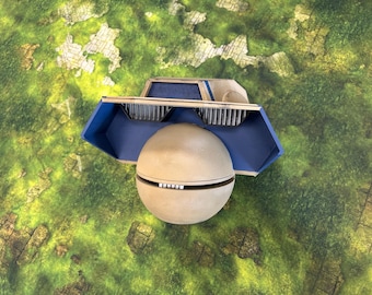 May include: A top-down view of a model spaceship with a spherical body and a blue and tan upper structure. The model is set against a green and brown terrain mat, suggesting a tabletop game setting.