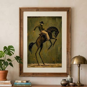 Könnte beinhalten: Ein gerahmter Kunstdruck mit einem Ölgemälde eines Cowboys mit schwarzem Hut und grünem Hemd auf einem sich aufbäumenden dunkelbraunen Pferd vor olivgrünem Hintergrund, in einem Holzrahmen mit weißem Passepartout.