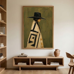 May include: A framed wall art print featuring a painting of a person in profile wearing a wide-brimmed black hat and a cream-colored blanket with bold black geometric patterns against a muted olive green background, displayed in a room.