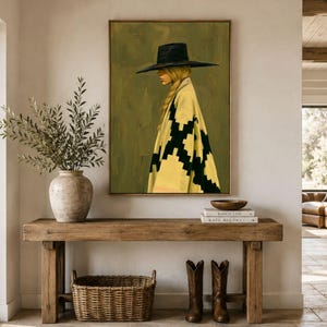 May include: A rectangular wall art print featuring a profile illustration of a person wearing a wide-brimmed black hat and a yellow poncho with a black geometric pattern against a muted olive green background, displayed in a thin wooden frame.