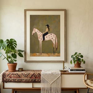 May include: A framed wall art print featuring a stylized illustration of a person wearing a black hat and dark clothing riding a light pink horse decorated with dark red star patterns, set against a muted olive green textured background.