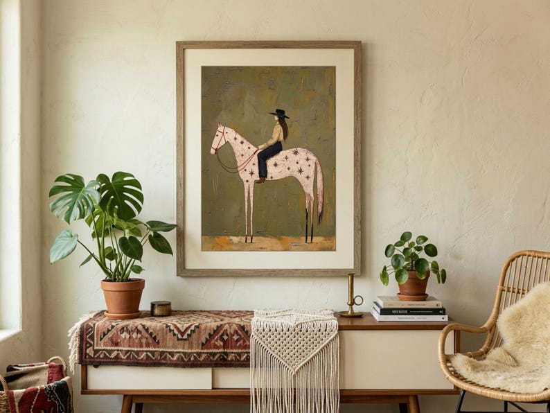 May include: A framed wall art print featuring a stylized illustration of a person wearing a black hat and dark clothing riding a light pink horse decorated with dark red star patterns, set against a muted olive green textured background.