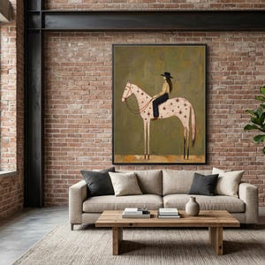 May include: A framed wall art print featuring a stylized illustration of a person wearing a black hat riding a light-colored horse with dark star patterns on its body, set against a muted olive green background with a textured paint effect.