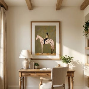 May include: A framed wall art print featuring a stylized illustration of a person wearing a black hat and dark clothing riding a light-colored horse with dark spots and star-shaped markings against a muted olive green background.